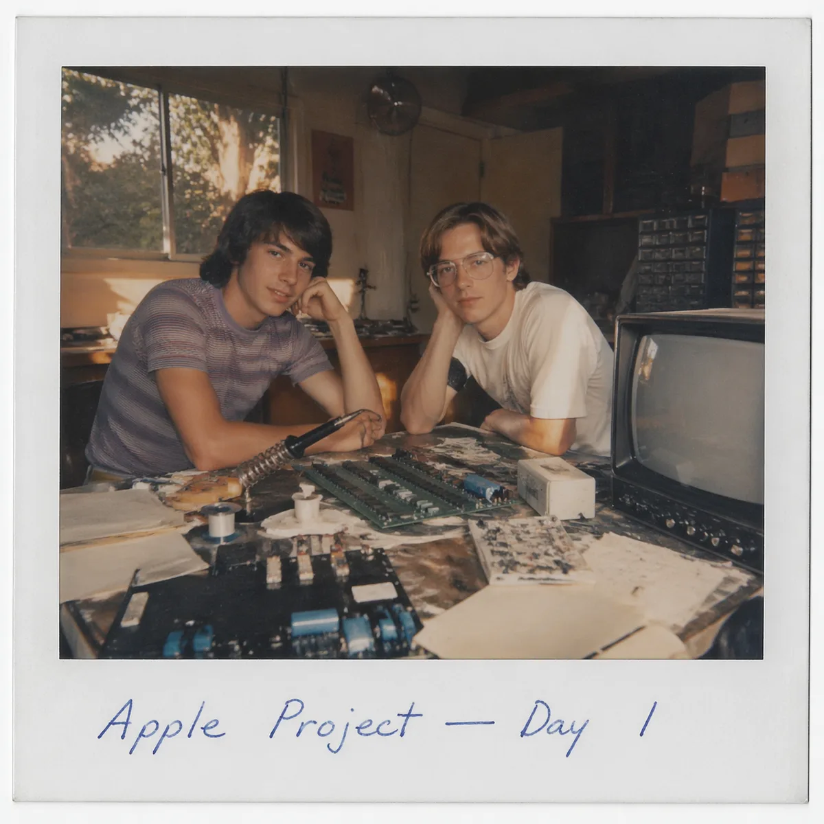 A hyperreal 1976 Polaroid of two teenage boys in a Cupertino garage, surrounded by circuit boards, a soldering iron and a CRT, late-afternoon sun streaming in, authentic white Polaroid border with handwritten caption in blue ballpoint 'Apple Project — Day 1'.